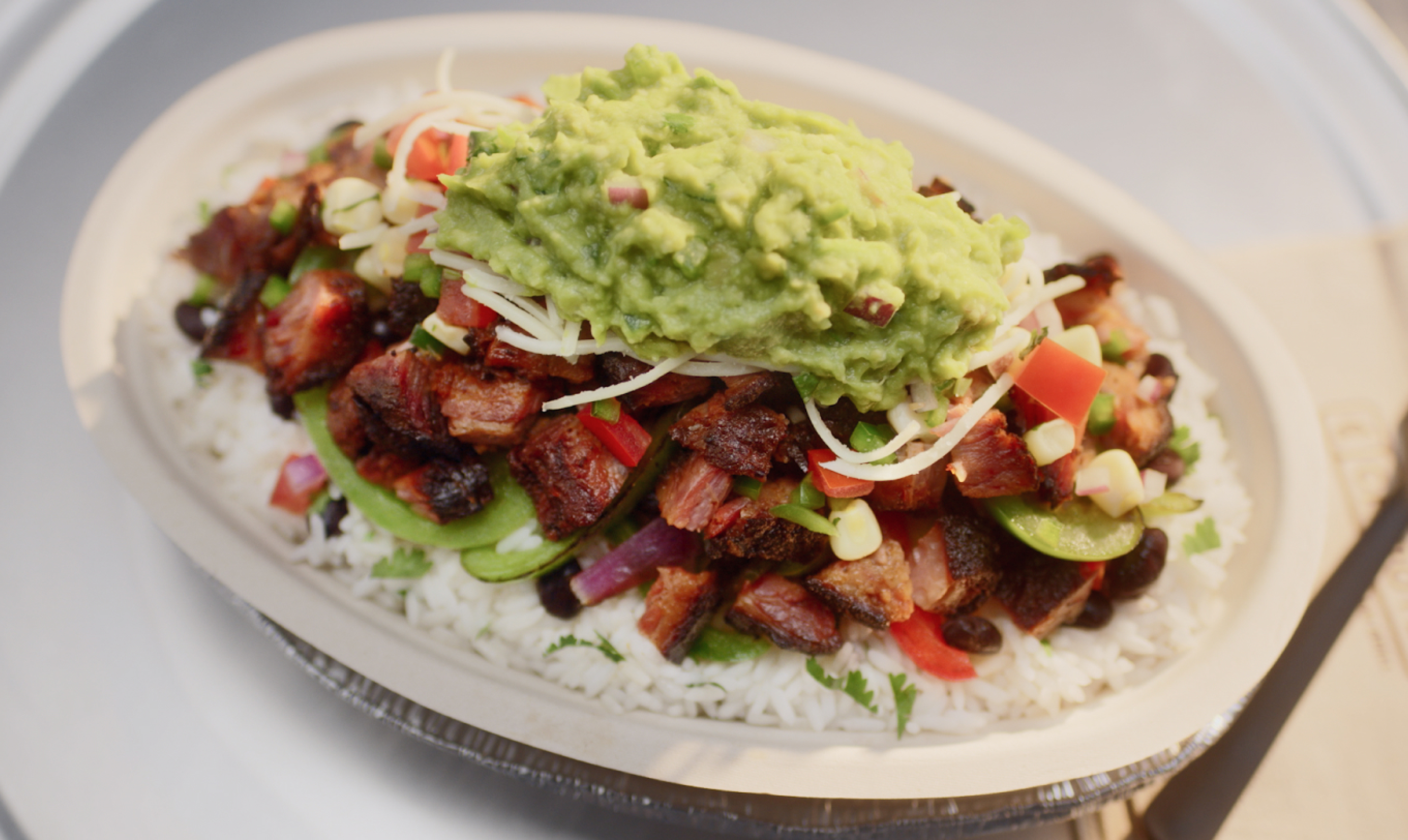 A Smoked Brisket Burrito Bowl with white rice, black beans, fajita veggies, hand-chopped Smoked Brisket, fresh tomato salsa, roasted chili corn salsa, monterey jack shredded cheese, and topped with hand-mashed guac.