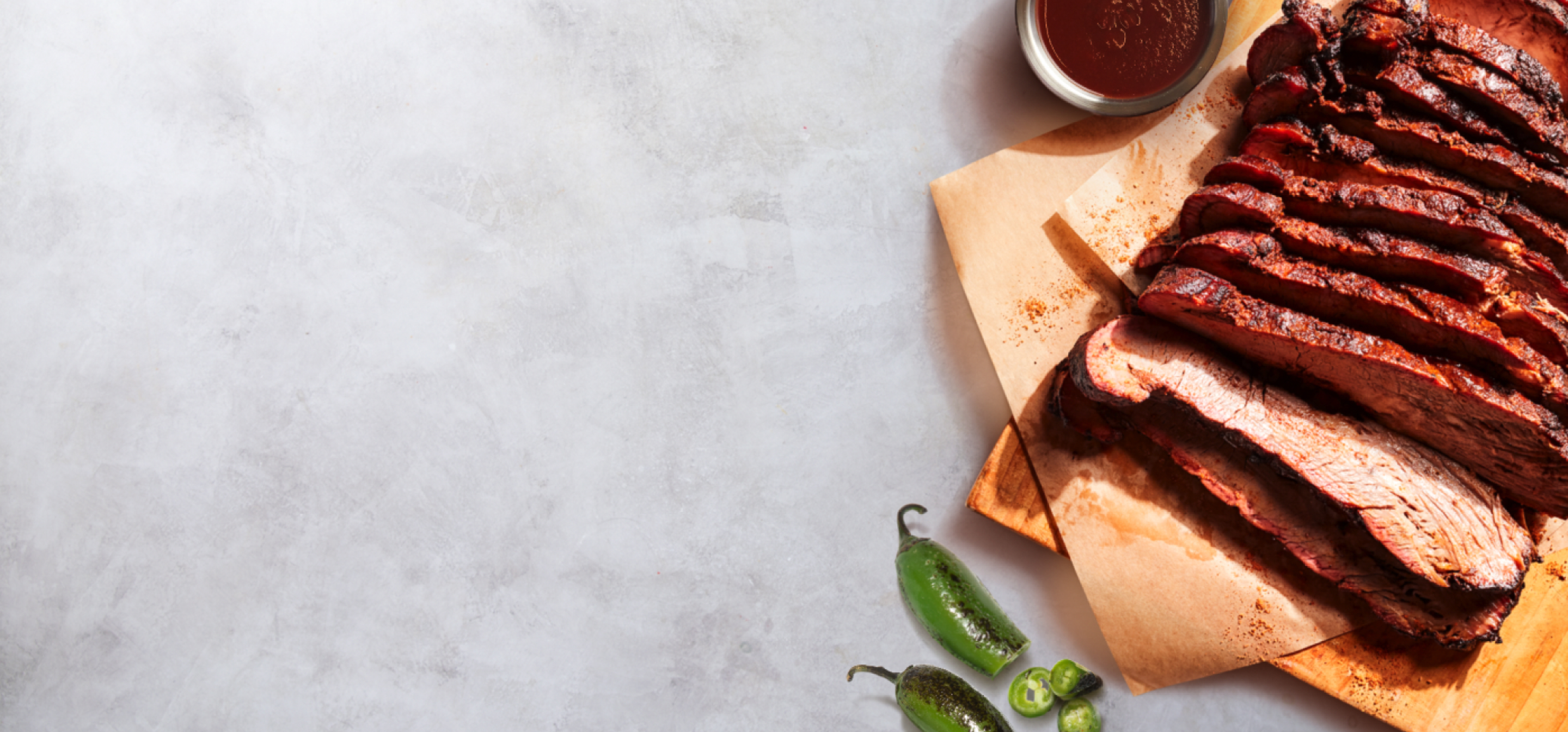 A wood cutting board of fresh sliced Smoked Brisket laying on a marble slab with a side of Chipotle’s signature sauce marinade and sliced jalapeño peppers.